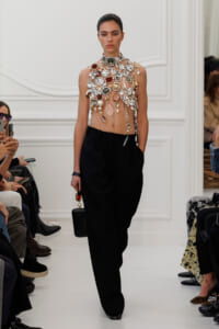 Model walks the runway wearing an elaborate gem-studded chest piece with black tailored trousers and a small black handbag on a white set. cool, high-fashion scene.