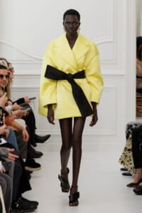 Model walks a runway in an oversized pale yellow coat with a wide black belt, audience seated along the sides.