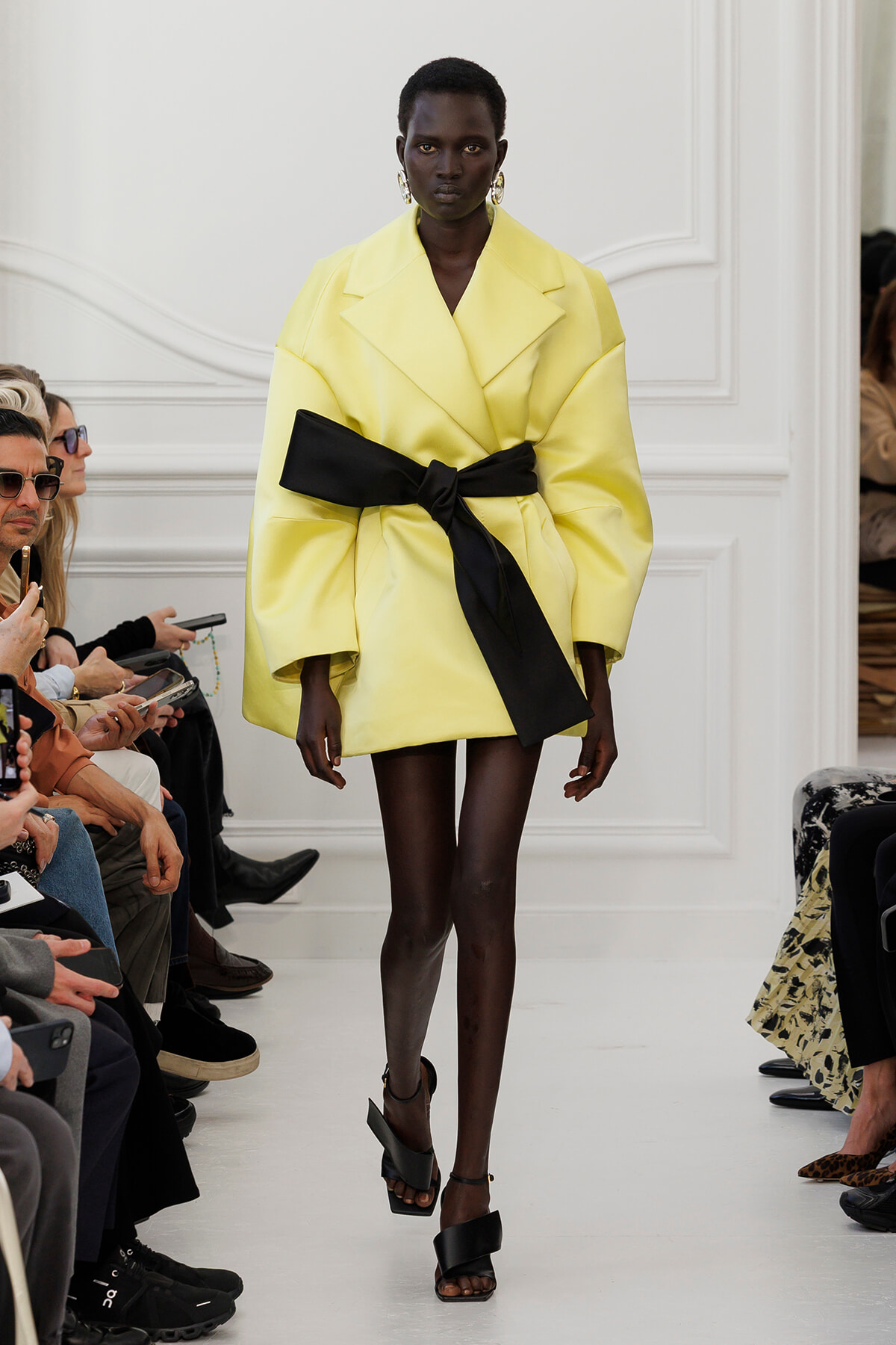 Model walks a runway in an oversized pale yellow coat with a wide black belt, audience seated along the sides.