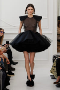 Model walking a runway in a sheer black bodice with sculpted shoulders and a voluminous layered tulle skirt, wearing large jeweled earrings.