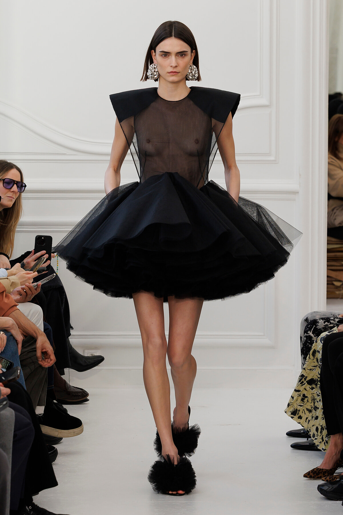 Model walking a runway in a sheer black bodice with sculpted shoulders and a voluminous layered tulle skirt, wearing large jeweled earrings.