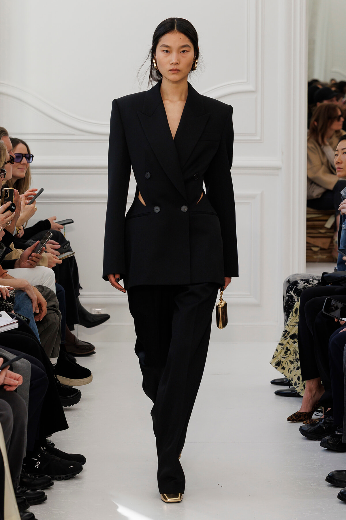 Model on a fashion-show runway wearing a structured black double-breasted blazer with side cutouts, black wide-leg pants, and gold accessories including a small handbag and square-toe shoes.