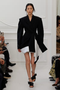 Model walking the runway in a black oversized blazer dress with a white asymmetric hem, large hoop earrings, and black sandals.