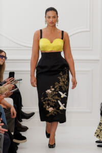 Fashion model walking a runway in a yellow textured bralette top and a black embroidered midi skirt with birds and flowers.