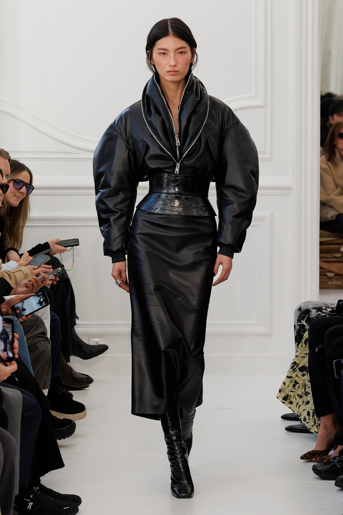 Model walks a runway in an all-black leather ensemble: oversized zip-front jacket cinched at the waist with a glossy belt, paired with a long skirt and knee-high boots, audience along the sides.