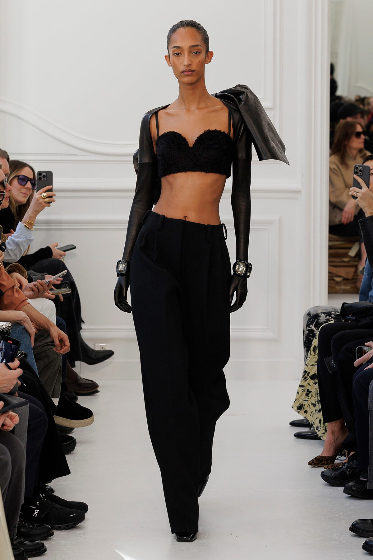 Runway model wears a black lace bralette, black gloves, and high-waisted black trousers with a leather jacket draped over her shoulders during a fashion show.