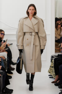 Model walks a runway in a beige oversized trench coat with wide lapels and a belt, wearing black gloves and glossy tall boots.