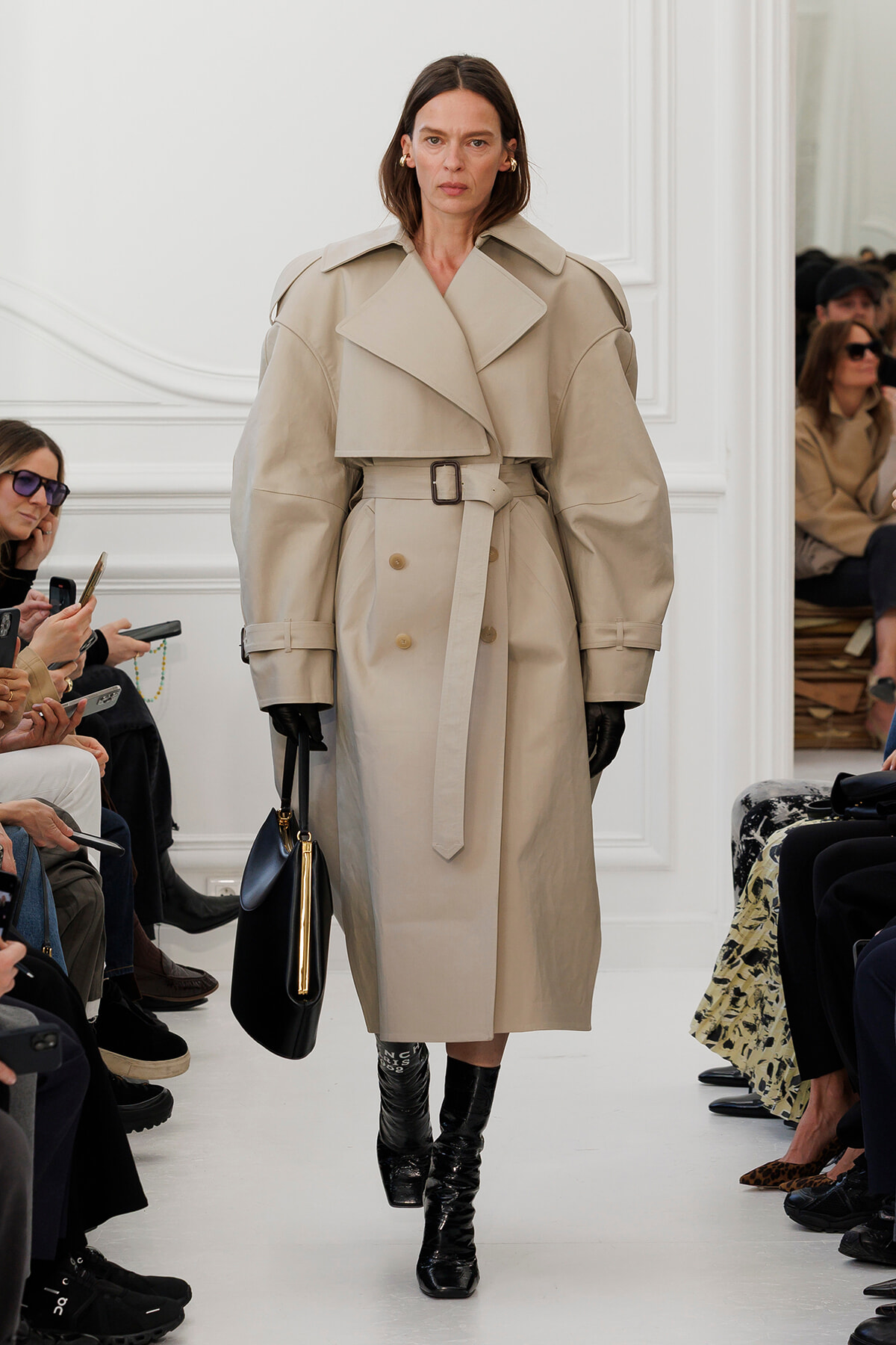 Model walks a runway in a beige oversized trench coat with wide lapels and a belt, wearing black gloves and glossy tall boots.