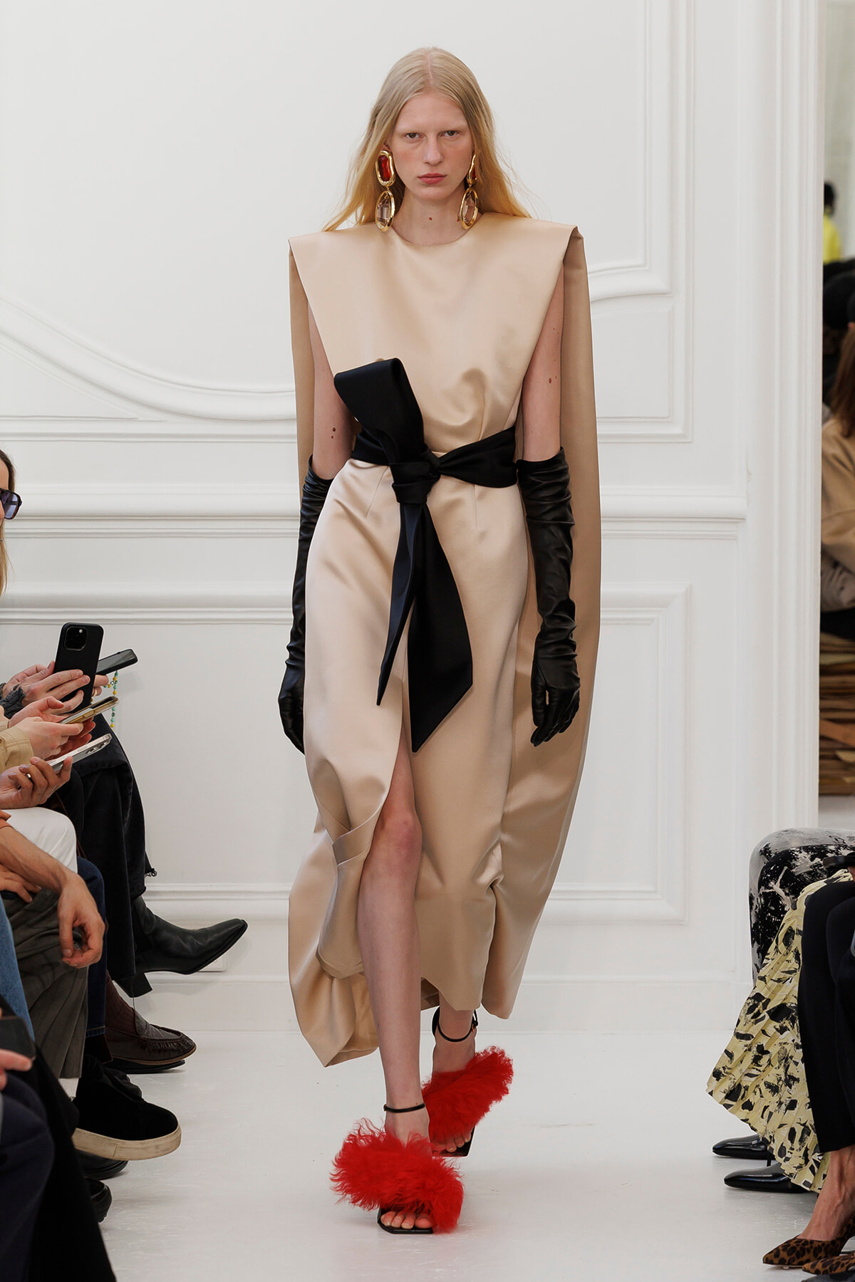 Model walking a runway in a satin beige dress with a black sash, long black gloves, and red feathered sandals during a fashion show.