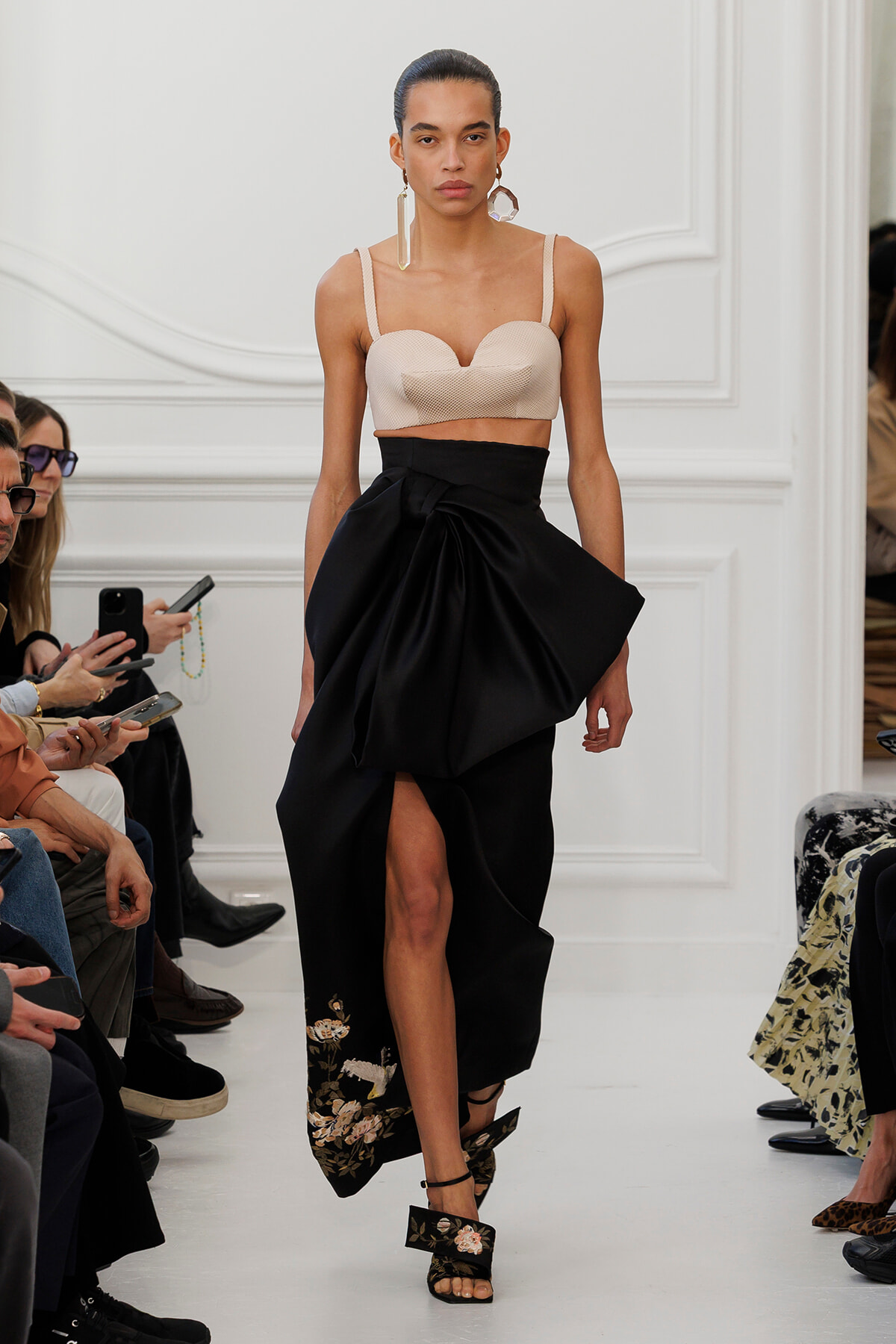 Model walking a runway in a beige strapless bustier top and a black draped, high-waisted skirt with floral embroidery at the hem. Audience visible on the left.