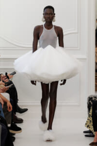 Black model walks a runway in a white halter-neck gown with a dramatic voluminous fluffy skirt and large statement earrings.