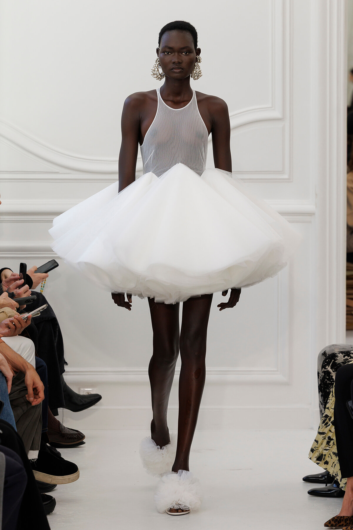 Black model walks a runway in a white halter-neck gown with a dramatic voluminous fluffy skirt and large statement earrings.