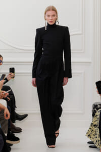Model on a runway wearing a structured black jumpsuit with a high neckline and wide-leg pants, accessorized with large geometric earrings and black heeled sandals.