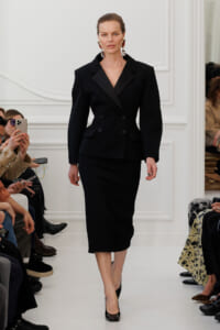 Model walking a runway in a tailored black double-breasted blazer and midi skirt, wearing bold dangling earrings.
