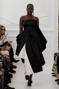 Model walking a runway in a strapless black gown with a sculptural bow and white underlayer, large dangling earrings visible.