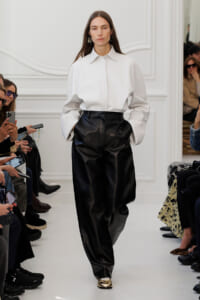 Runway look: model in oversized white shirt and black leather wide-leg pants, walking a white catwalk with seated audience on both sides.