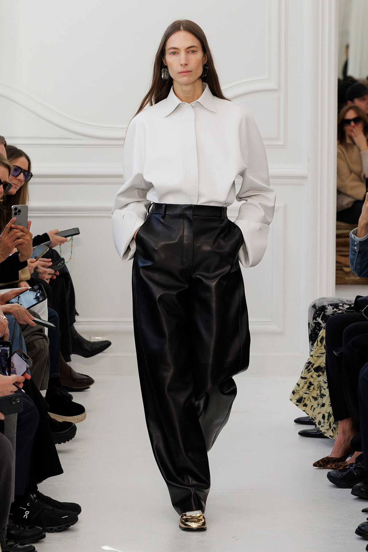Runway look: model in oversized white shirt and black leather wide-leg pants, walking a white catwalk with seated audience on both sides.