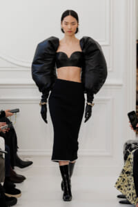 Model on a runway wearing a black two-piece ensemble: a structured cropped top with heart-shaped neckline, dramatic oversized puff sleeves, and a sleek midi skirt with patent boots.