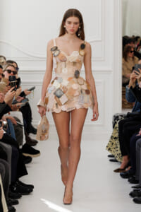 Runway model wearing a beige patchwork mini dress made from circular and square fabric patches, with nude heels, walking the catwalk.