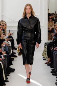 Model walking a white runway in a black leather dress with voluminous sleeves, holding a gold clutch.