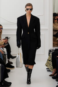 Model walking a white runway in an oversized black blazer-style dress with a single gold button, bold shoulders, black gloves, oversized sunglasses, and a white handbag with gold accents.