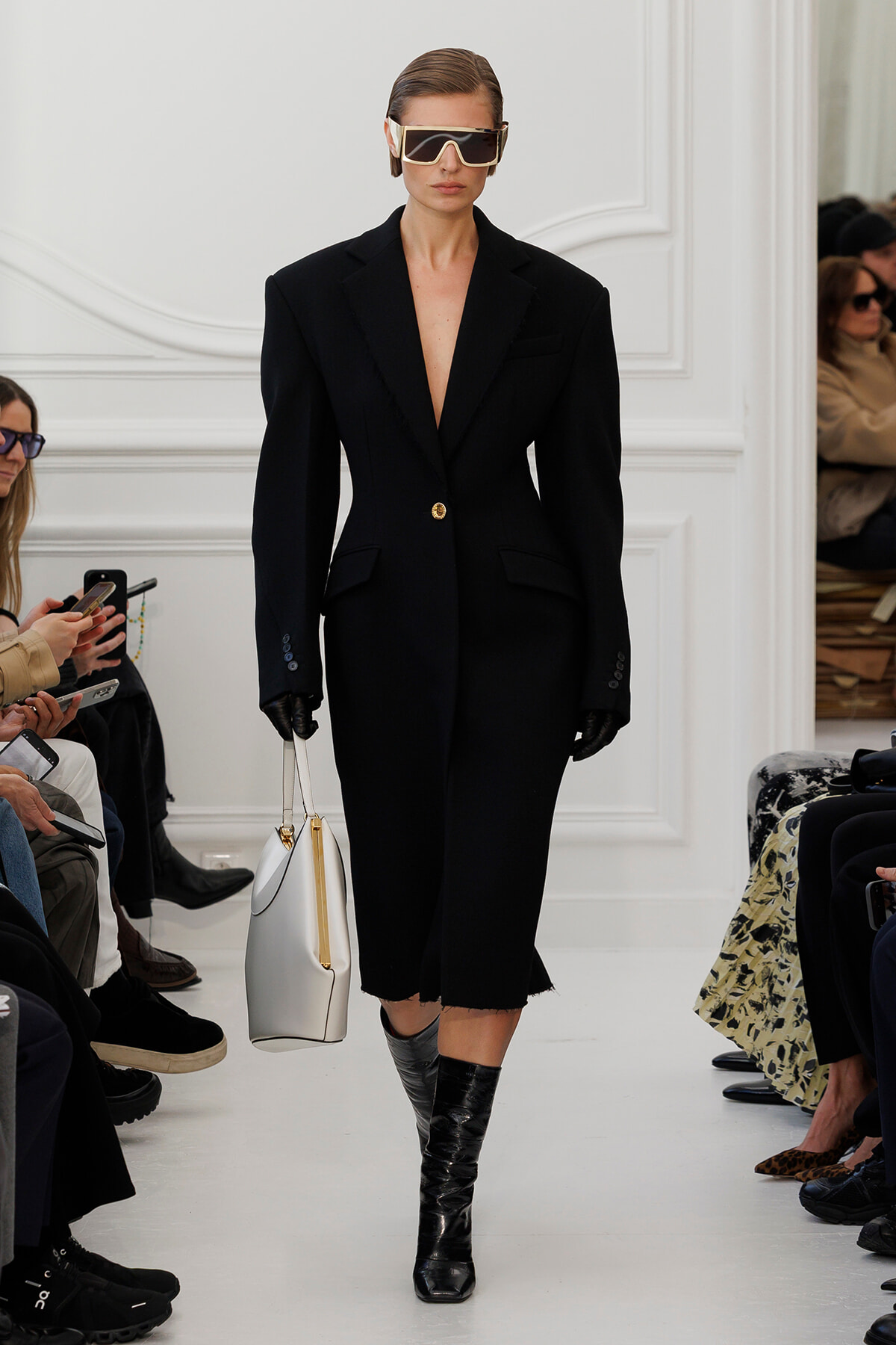 Model walking a white runway in an oversized black blazer-style dress with a single gold button, bold shoulders, black gloves, oversized sunglasses, and a white handbag with gold accents.