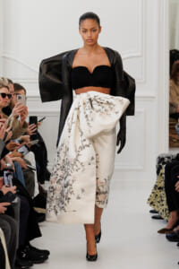 Model walking the runway in a black textured strapless top, oversized cape sleeves, and a white floral-embroidered draped wrap skirt; audience with phones in background.