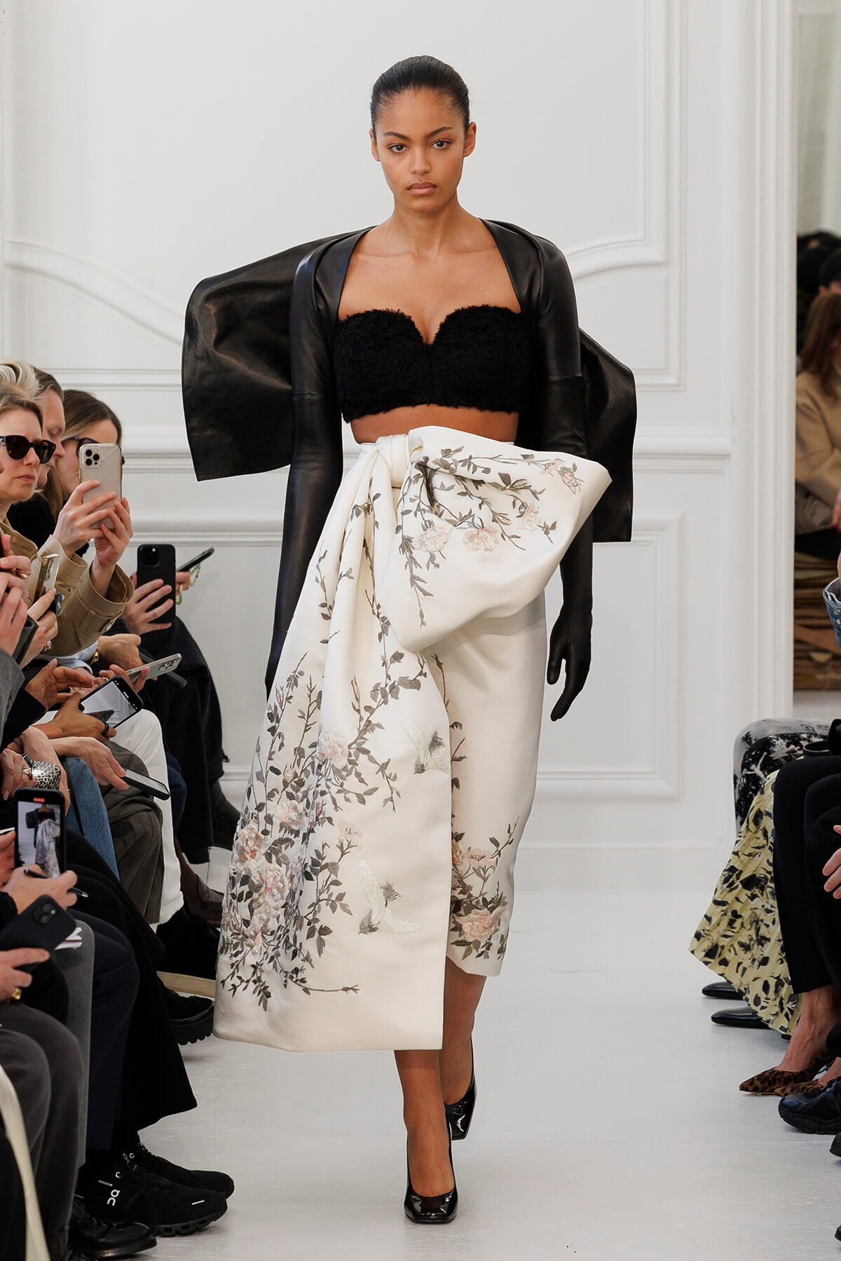 Model walking the runway in a black textured strapless top, oversized cape sleeves, and a white floral-embroidered draped wrap skirt; audience with phones in background.