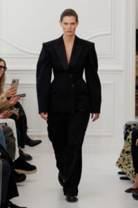 Model in an all-black tailored suit with a bold-shouldered blazer and wide-leg pants walking the runway, audience taking photos on the sides.