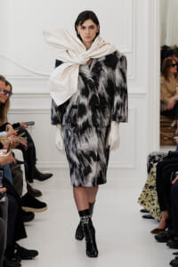 Model walks a runway in a black-and-white abstract coat, an oversized cream scarf knotted at the neck, white gloves, and black boots with white text.