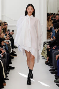Model walking a fashion runway in a loose white oversized shirt-dress with a dramatic draped shoulder, paired with black ankle boots; audience seated along the sides watching and filming.