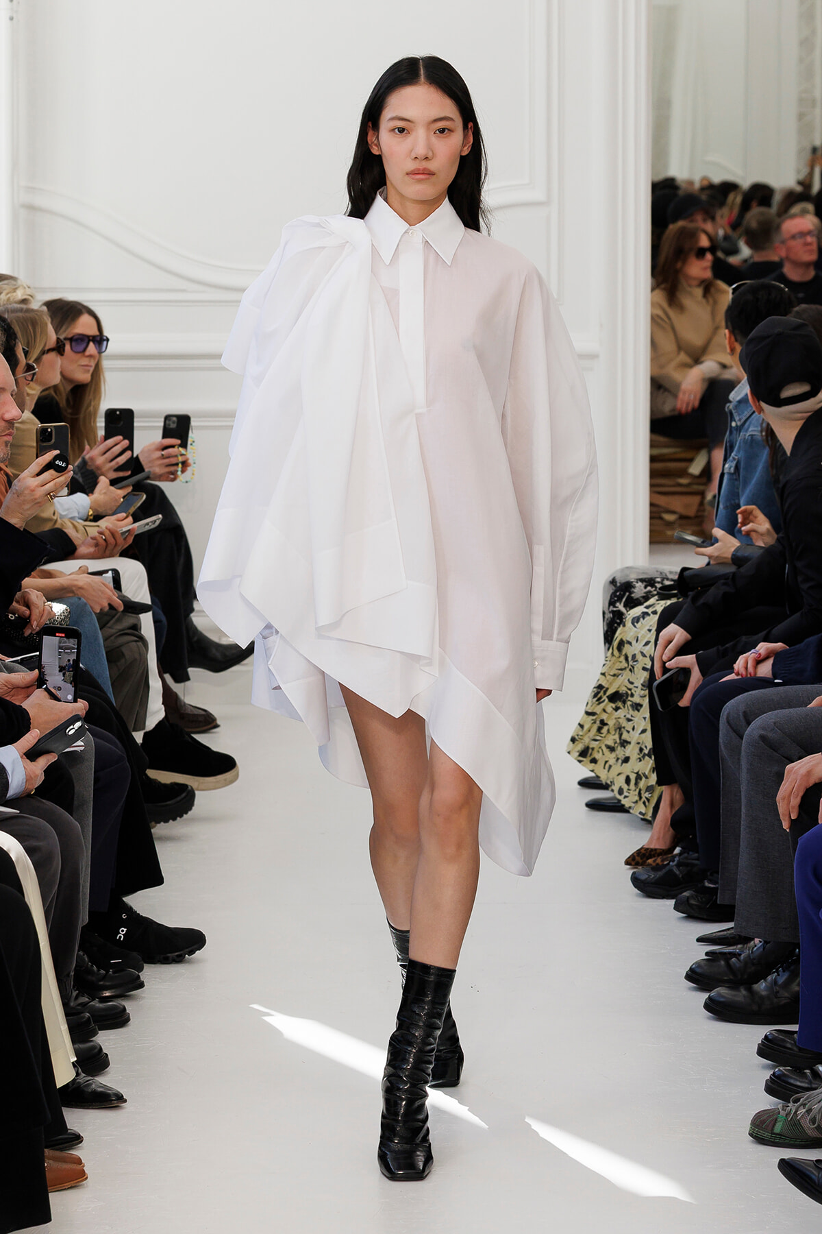 Model walking a fashion runway in a loose white oversized shirt-dress with a dramatic draped shoulder, paired with black ankle boots; audience seated along the sides watching and filming.