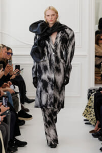 Model walks the runway in a black-and-white abstract-patterned coat with a large black scarf wrapped around her neck, wearing black gloves.