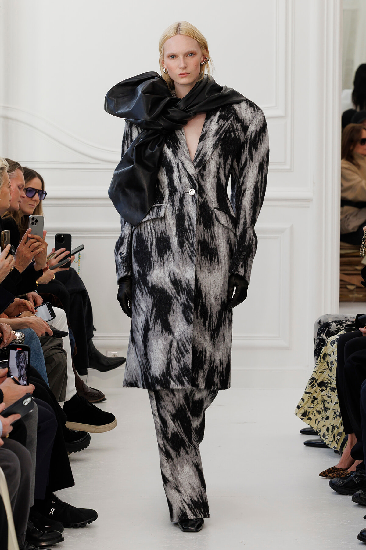 Model walks the runway in a black-and-white abstract-patterned coat with a large black scarf wrapped around her neck, wearing black gloves.