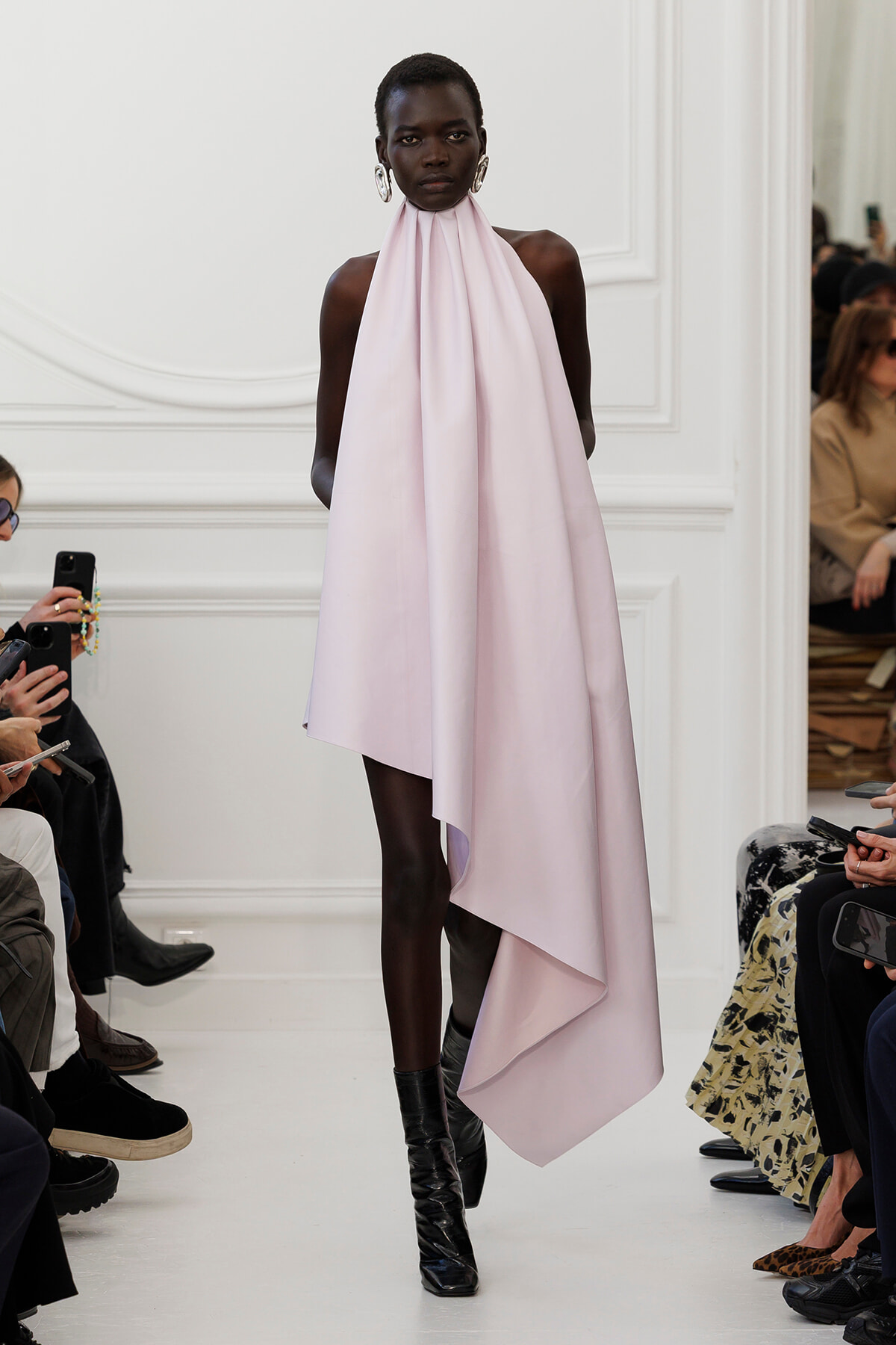 Runway model in a pale pink halter-neck gown with a long draped cape-like hem.