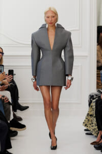 Model walks a fashion show in a gray structured mini dress with a plunging V neckline and broad shoulders, paired with black heels and oversized silver watches.