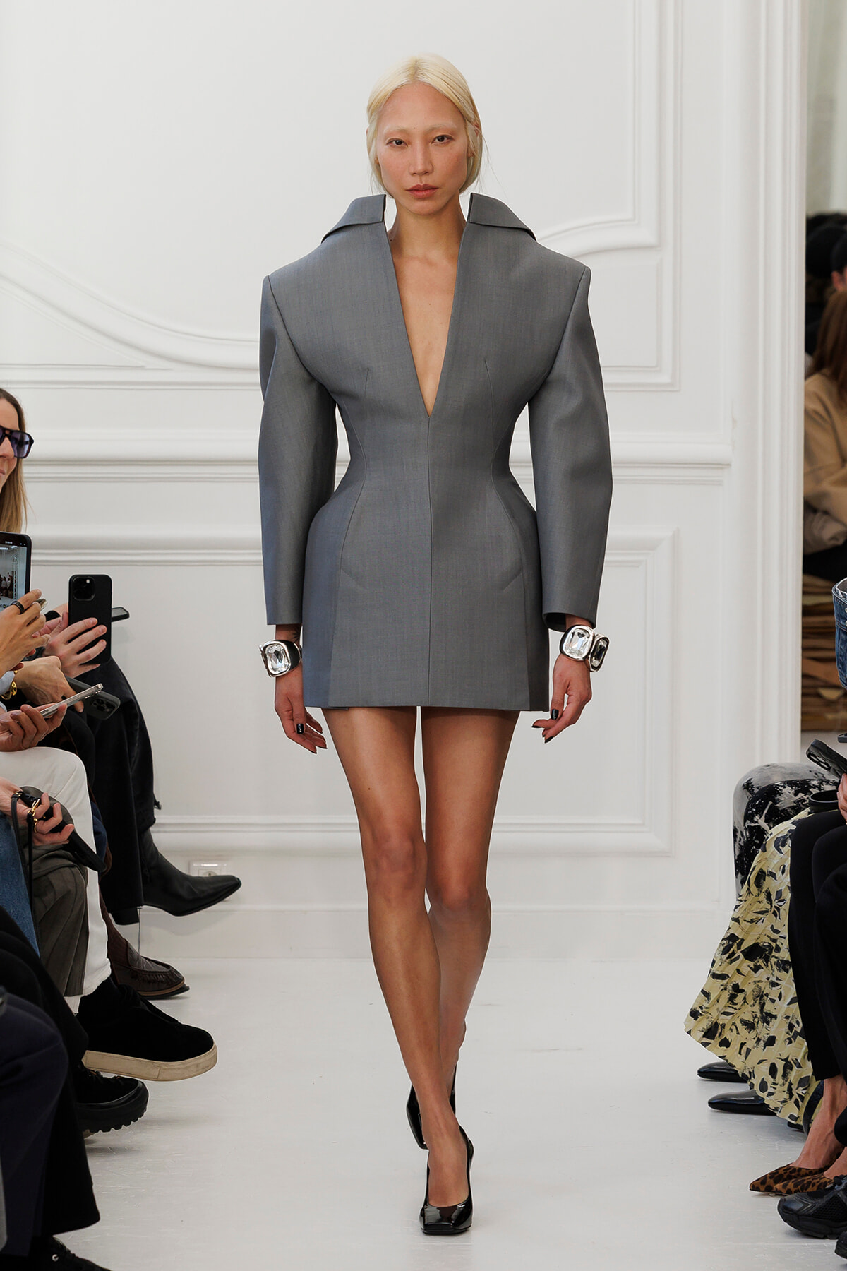 Model walks a fashion show in a gray structured mini dress with a plunging V neckline and broad shoulders, paired with black heels and oversized silver watches.