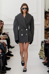 Model walks a runway in a large gray double-breasted wool coat, black gloves, and oversized sunglasses.