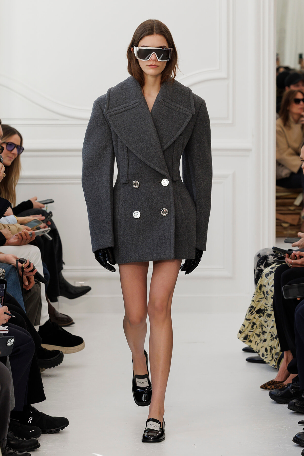 Model walks a runway in a large gray double-breasted wool coat, black gloves, and oversized sunglasses.