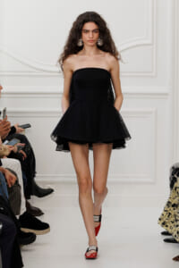Model on a runway wearing a strapless black lace mini dress with voluminous skirt and large chandelier earrings, audience seated nearby.