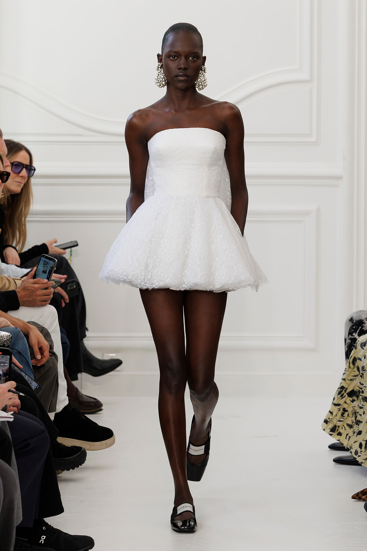 Model walks a runway in a strapless white lace mini dress with a voluminous, bell-shaped skirt and large pearl earrings.