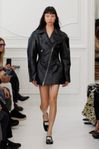 Model walks the runway in a black oversized leather biker jacket and black patent loafers, audience visible on sides.
