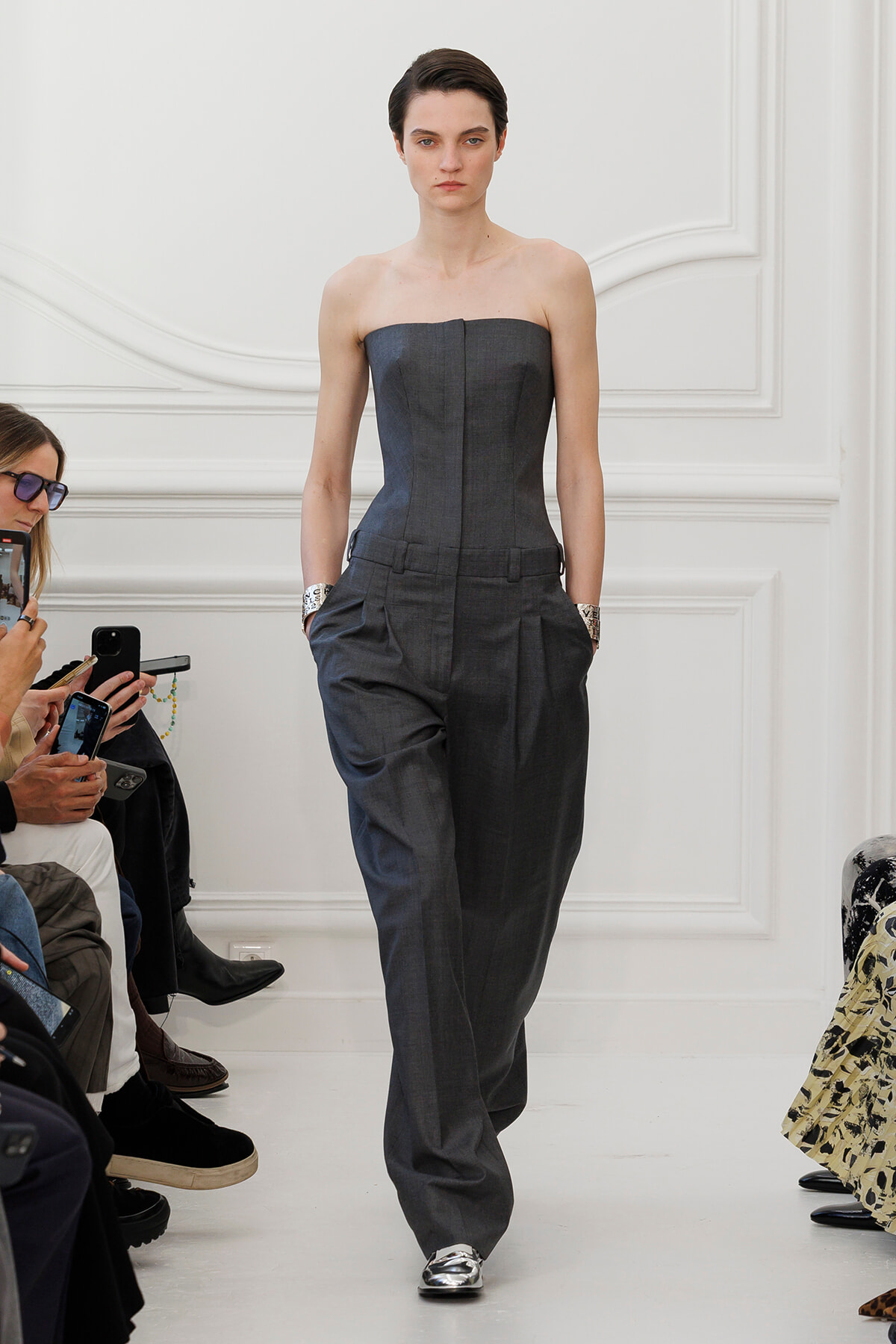 Model walks a runway in a strapless charcoal gray jumpsuit with wide legs; hands in pockets, metallic loafers visible.