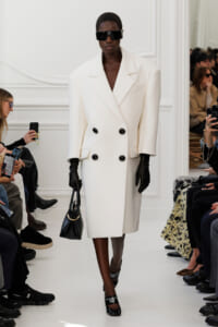 Model on a runway wearing an oversized white double-breasted coat, black gloves, sunglasses, and carrying a black handbag.