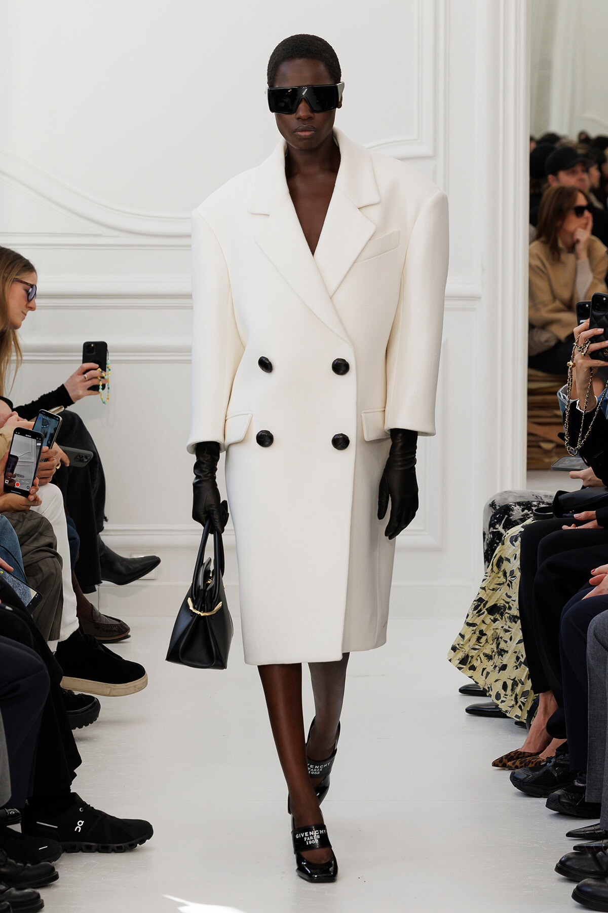 Model on a runway wearing an oversized white double-breasted coat, black gloves, sunglasses, and carrying a black handbag.
