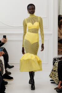 Model on a white runway wearing a bright yellow mesh dress with a high neck, long sleeves, and a voluminous ruffled hem paired with black boots