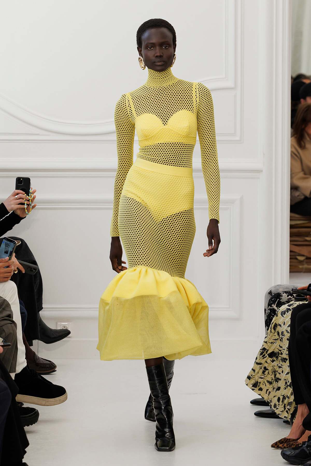 Model on a white runway wearing a bright yellow mesh dress with a high neck, long sleeves, and a voluminous ruffled hem paired with black boots