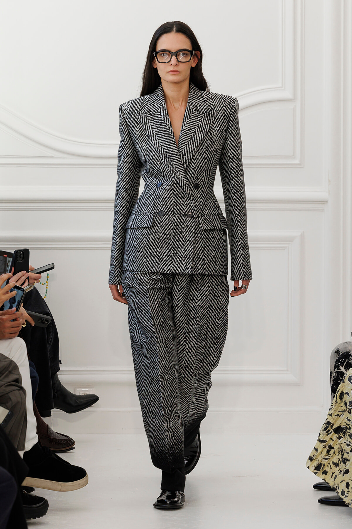 Model walking the runway in a black-and-white herringbone two-piece suit with oversized shoulders and glasses.
