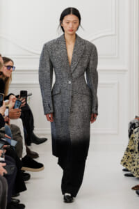 Model walking a runway in a gray-to-black herringbone coat over wide-legged black pants, audience filming with phones nearby.