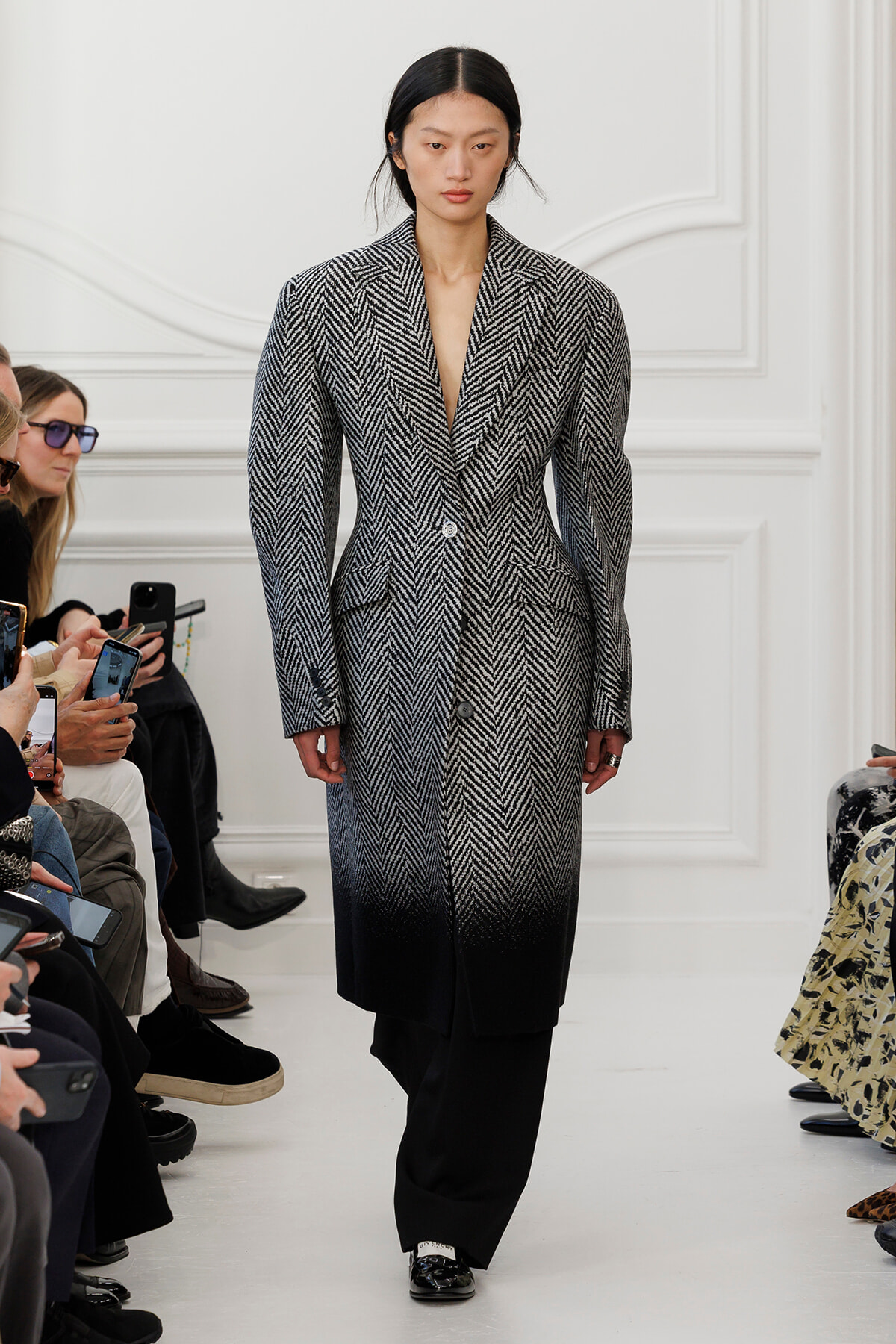 Model walking a runway in a gray-to-black herringbone coat over wide-legged black pants, audience filming with phones nearby.